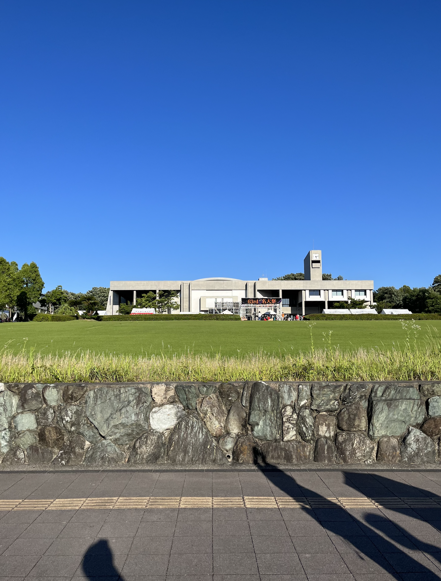 名古屋大学の豊田講堂の写真。坂になっている芝生の上に横長の灰色の建物が立っている晴れた日の写真。